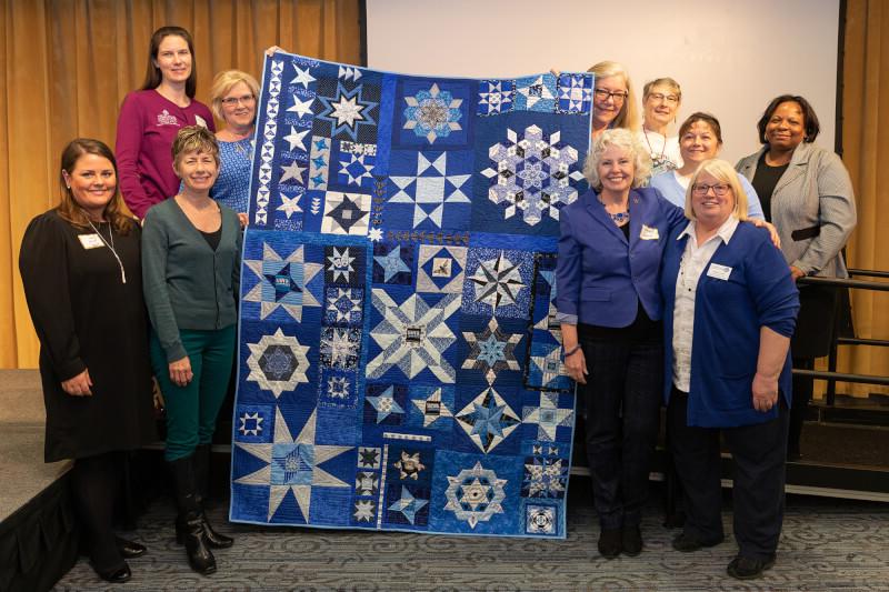 women standing in two rows surrounding a quilt