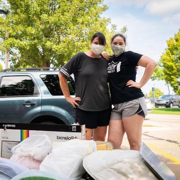 Two people stand with their arms around each other, posing behind a pile of someone's belongings.